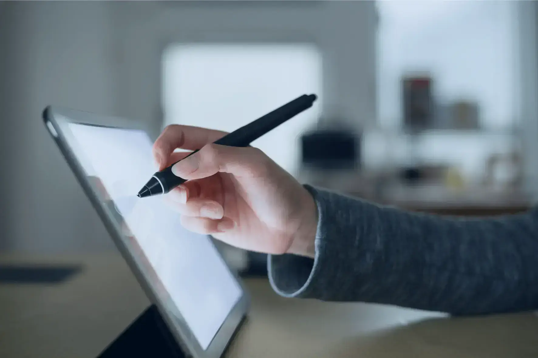 A close-up of a hand holding a black stylus pen and using it on a digital tablet in a professional workspace.